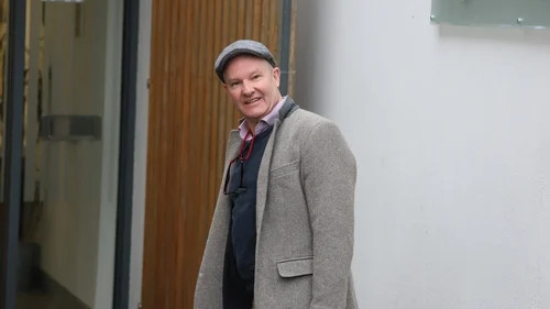 Smiling man in a gray coat and cap standing outside a building near a wooden door.