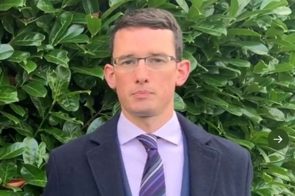 Man in a suit and striped tie standing outdoors, framed by dense green leaves.