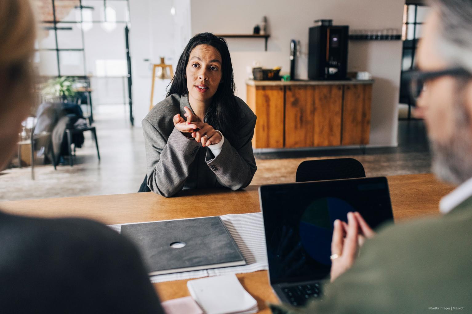 Woman in blazer leans forward at meeting table, gesturing while speaking to colleagues in office.