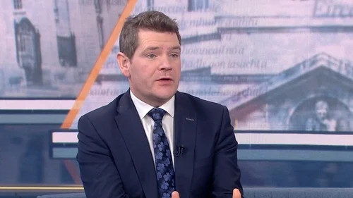 Man in a dark suit and tie speaking on a news studio set with city backdrop.