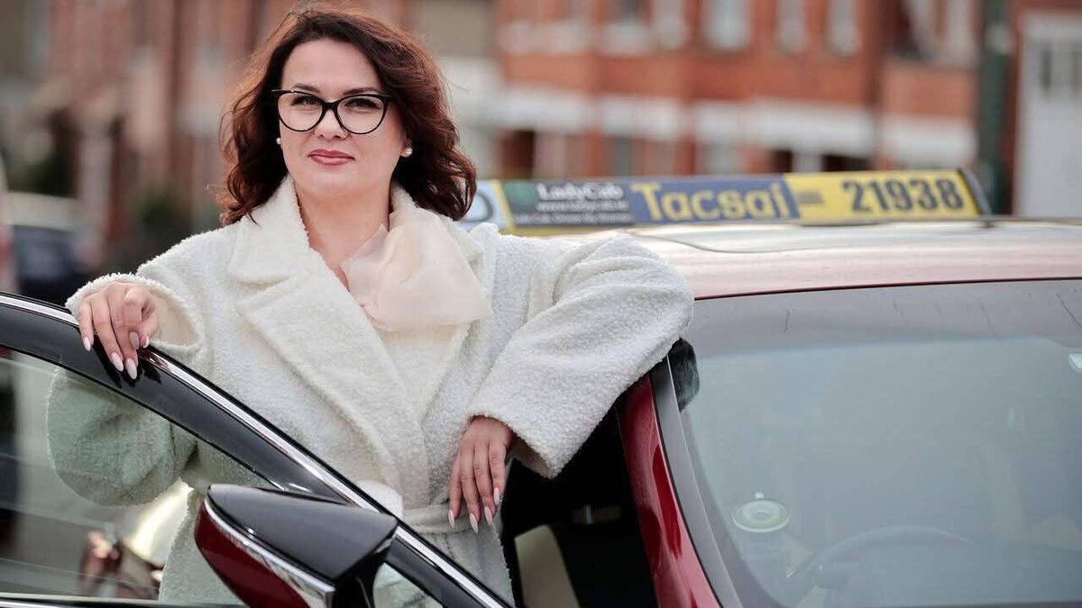 Woman with glasses in a white coat leans on a car door in front of brick buildings.
