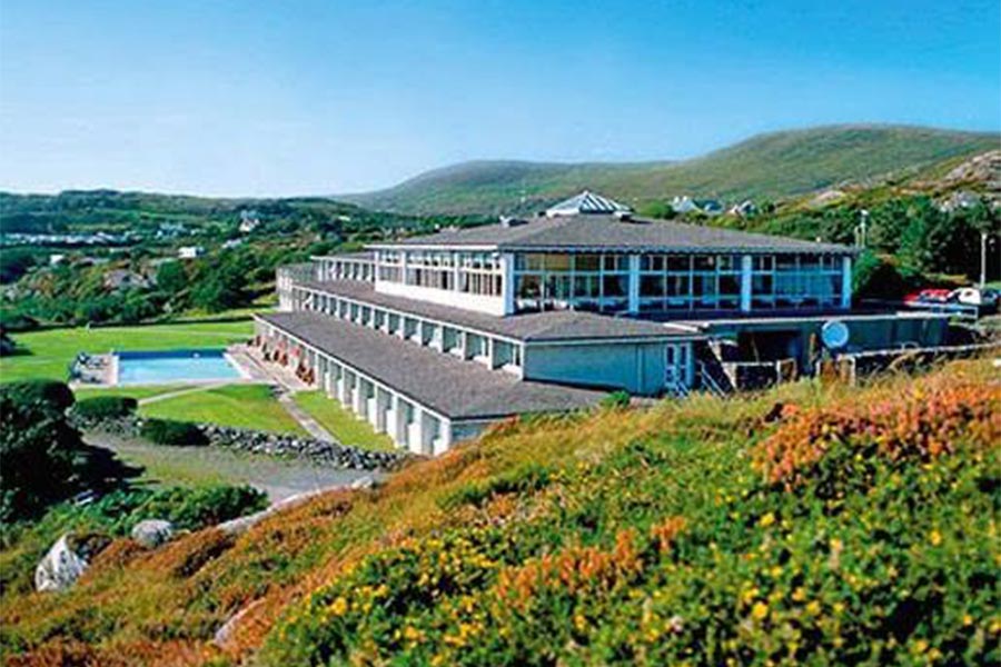 Modern resort building with blue windows, surrounded by green hills, gardens, and a swimming pool.