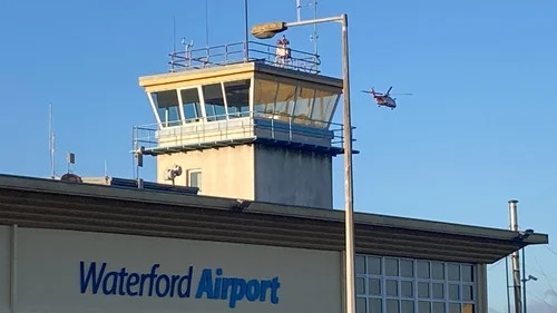 Waterford Airport control tower with blue sign, clear sky, and a small airplane flying overhead.
