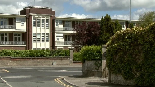 School buildings with brick facade and windows, roadside curve, hedges and trees under cloudy sky.