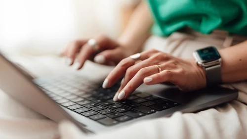 Hands typing on a laptop keyboard while wearing a smartwatch, with another hand resting nearby.