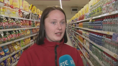 Woman in a grocery aisle between shelves of packaged food, speaking into a microphone.