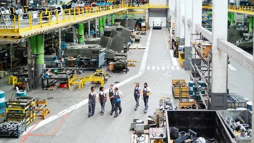 Workers walk through a large industrial factory floor with machinery, pipes, and overhead conveyor lines.
