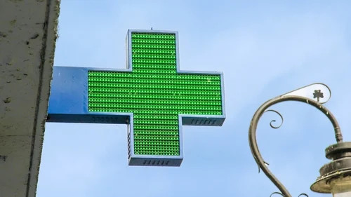 Green illuminated cross sign on building facade against blue sky, with decorative streetlamp nearby.