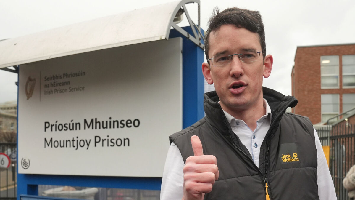Man in glasses and jacket gestures in front of Mountjoy Prison sign outside prison building.