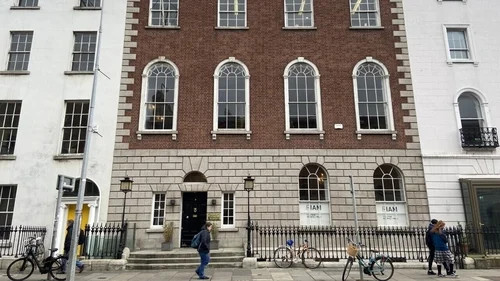 Brick and stone building facade with arched windows, pedestrians walking, and bicycles parked along the sidewalk.