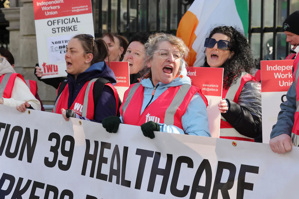 Protesters in red vests shout behind a banner reading “HEALTHCARE” during an official strike demonstration.