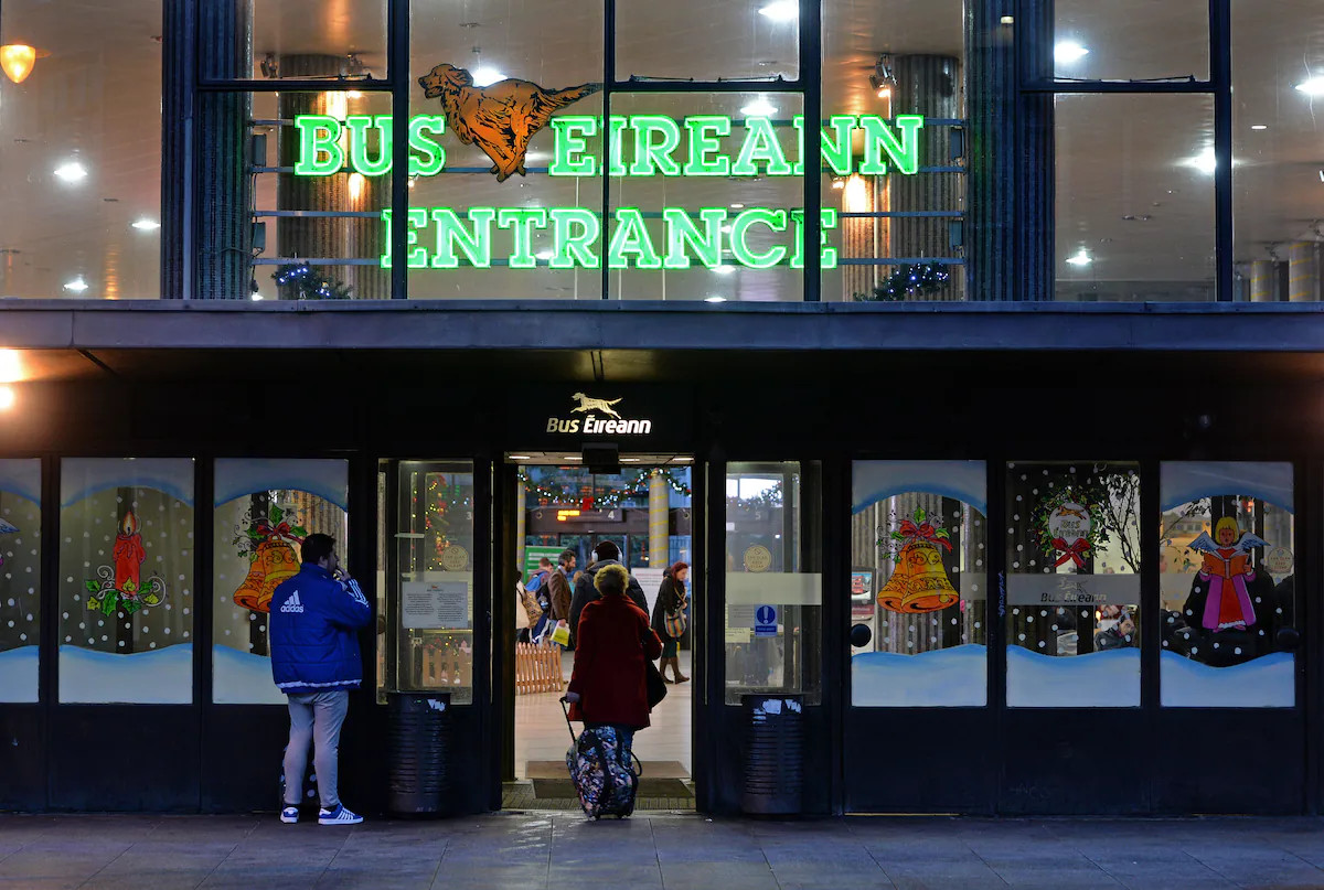People enter a bus station entrance with illuminated green sign reading “Bus Eireann” and “Entrance.”