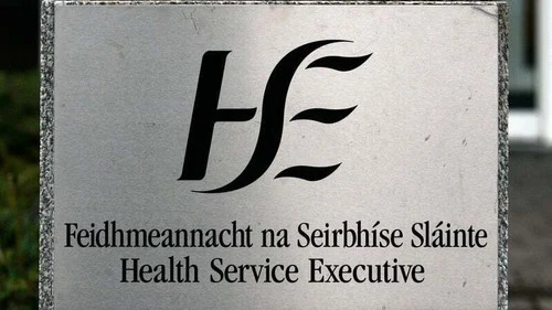 Metal sign with Health Service Executive text and stylized HSE logo, outdoors against a blurred background.