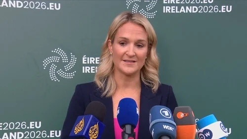 Blonde woman speaking at a press conference behind multiple microphones against an Ireland 2026 backdrop.