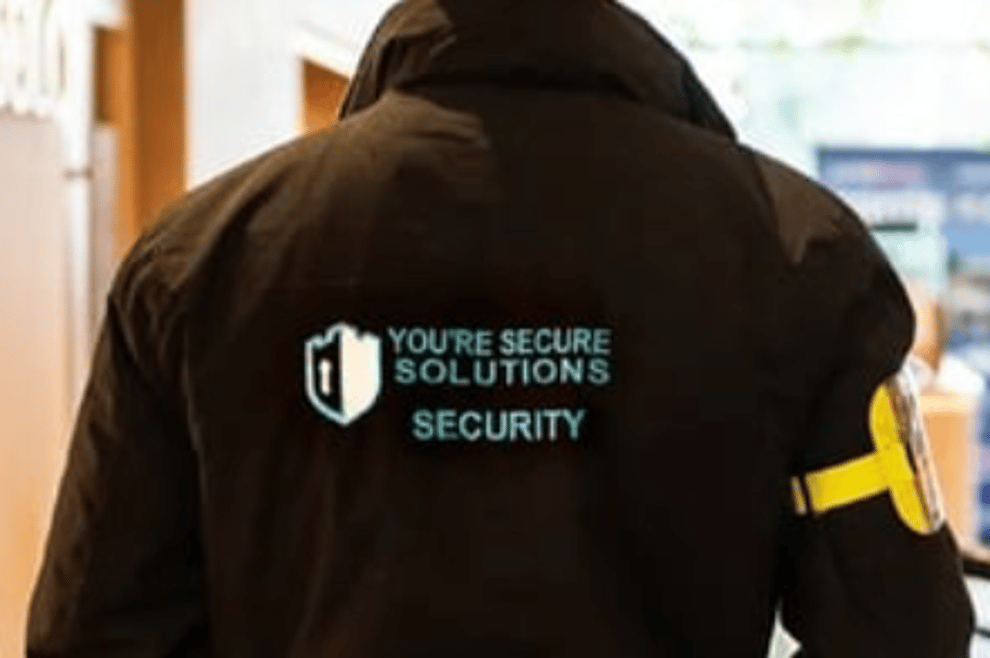 Security guard in dark jacket with “YOUR SECURE SOLUTIONS SECURITY” logo, standing indoors near windows.