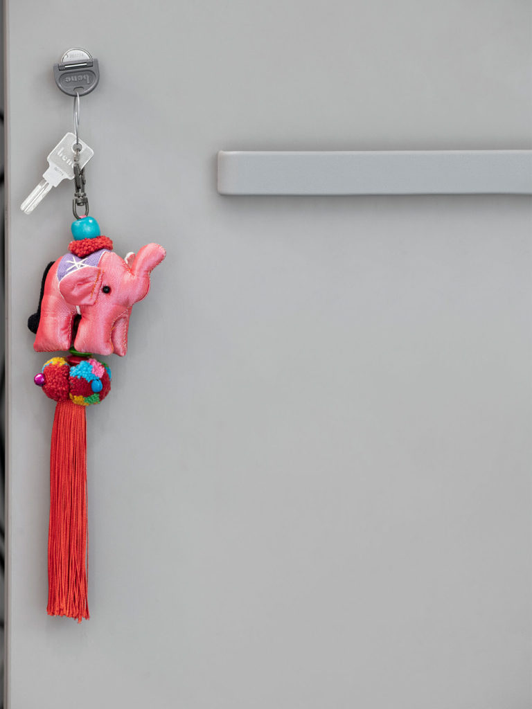 Pink plush elephant keychain with tassel and pom-poms hanging from a metal lock on a door.