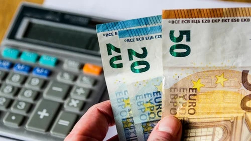 Hand holds euro banknotes and coins beside a calculator, suggesting budgeting or financial calculations.