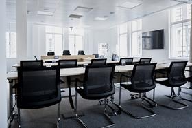 Bright modern conference room with long table, black chairs, and large windows under white ceiling lights.