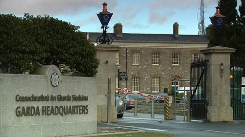 Garda Headquarters entrance with stone sign, iron gates, parked cars, and a cloudy sky background.