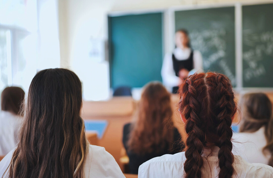 Woman with braided red hair sits in classroom, facing blurred teacher and students during presentation.