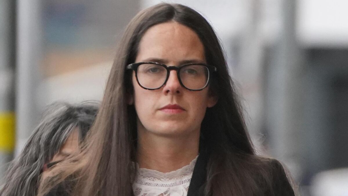 Woman with long hair and black glasses, serious expression, standing indoors with blurred background.