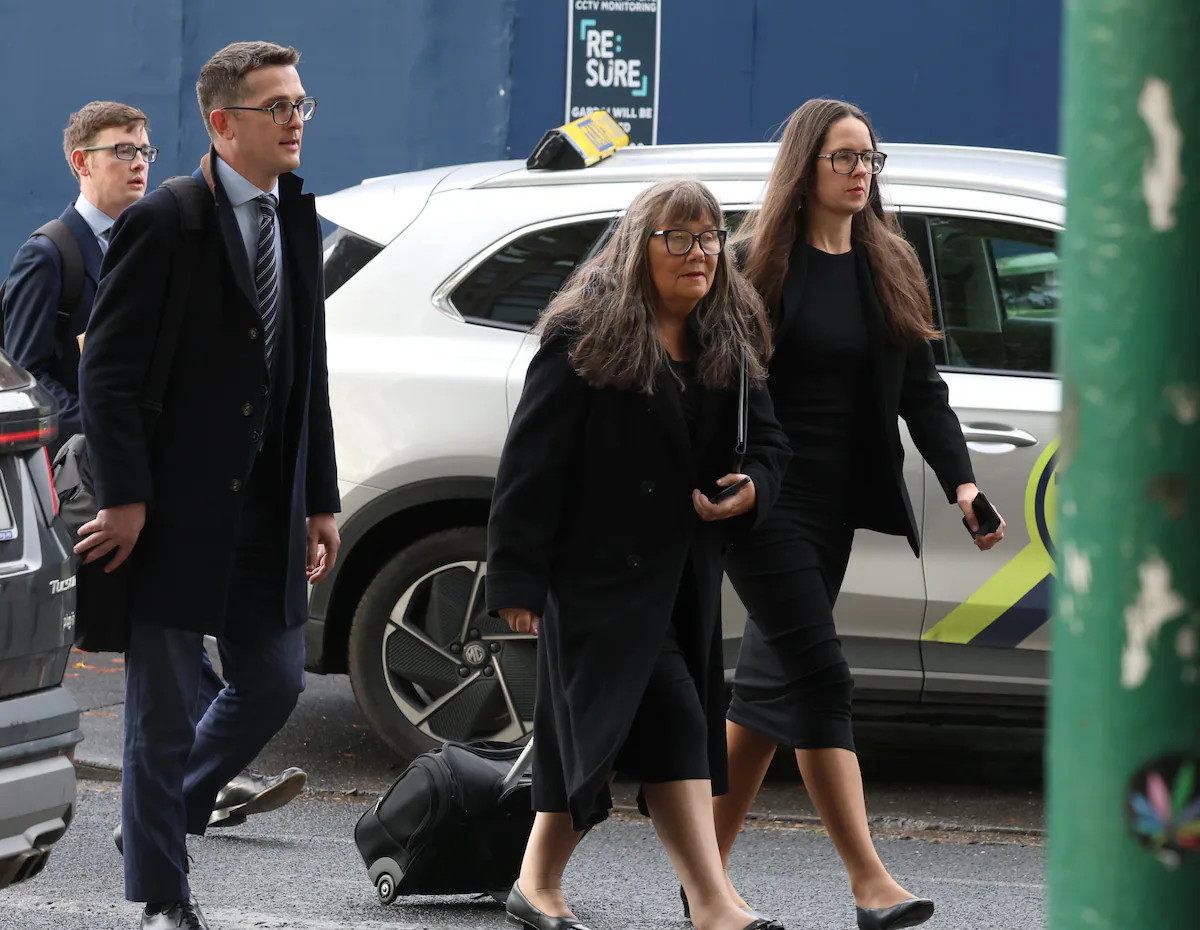 Three businesspeople in suits walk past a white SUV, one holding a phone, outside a building.