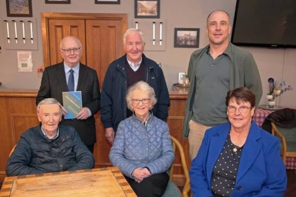 Five older adults pose indoors, three standing and two seated, smiling in a community room.