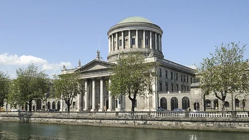 Historic neoclassical building with domed rotunda and columns beside a river, trees in foreground.